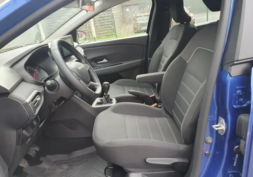 Vue intérieure côté conducteur d'une Dacia Sandero bleu Iron 2025, avec sièges noirs et accoudoir central.