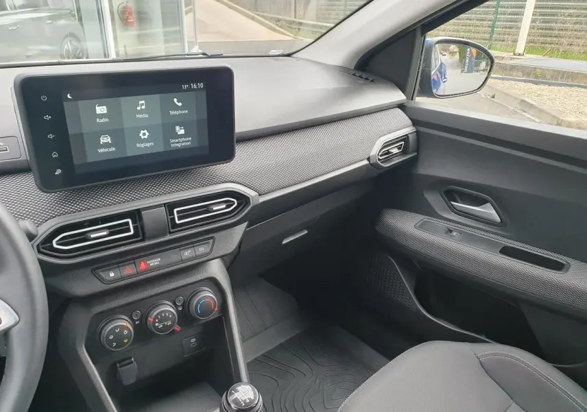 Vue intérieure côté passager du tableau de bord et console centrale de la Dacia Sandero 2025 avec écran tactile et commandes climatisation.