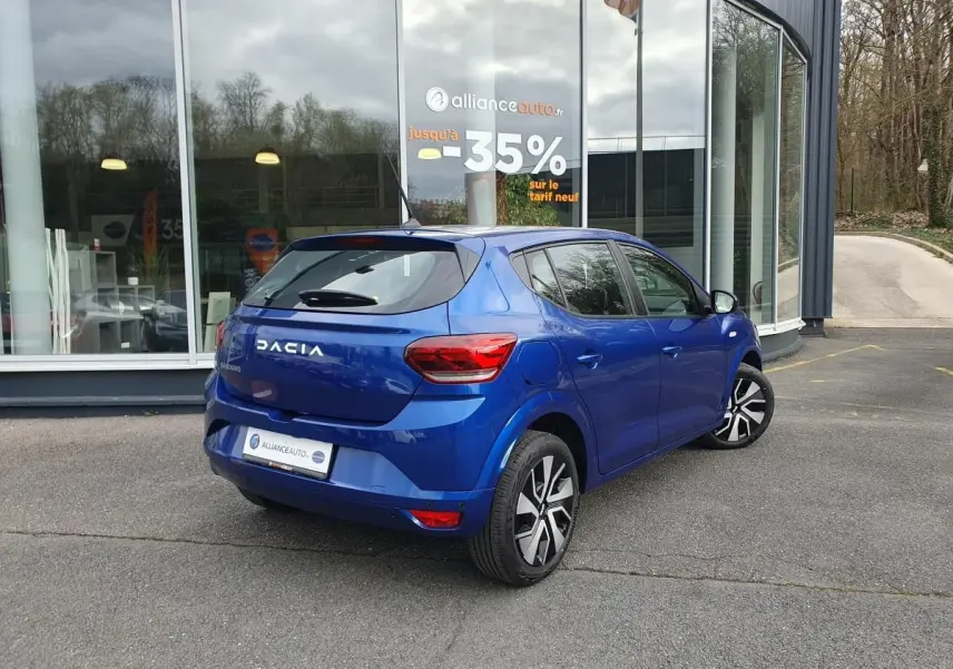Vue 3/4 arrière droite d'une Dacia Sandero bleu Iron avec enjoliveurs 16'' Atara, stationnée devant un bâtiment vitré.
