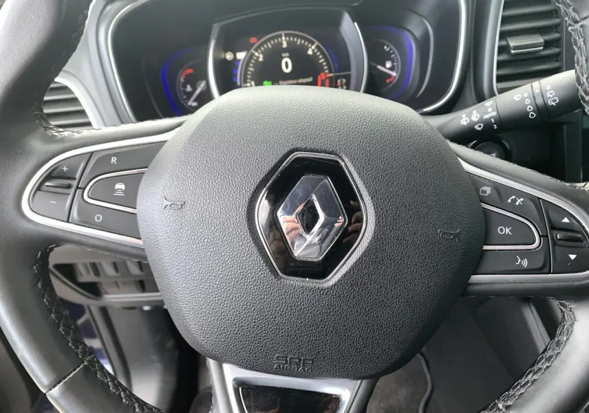 Vue rapprochée du volant en cuir noir avec logo Renault, tableau de bord digital bleu et commandes intégrées visibles.