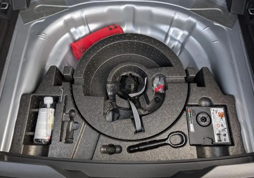 Vue plongeante sur le coffre ouvert d’un Volkswagen T-Roc gris, montrant la roue de secours et les outils de bord.