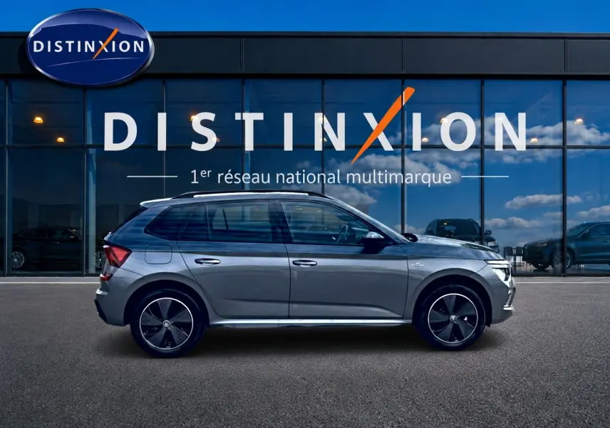 Vue de profil droit du Skoda Kamiq gris graphite avec jantes alu noires et barres de toit visibles devant une vitrine Distinxion.