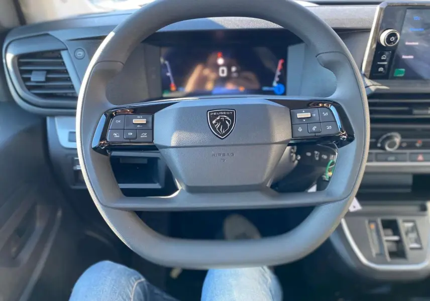 Vue rapprochée du volant Peugeot gris avec commandes intégrées et tableau de bord numérique flou en arrière-plan.