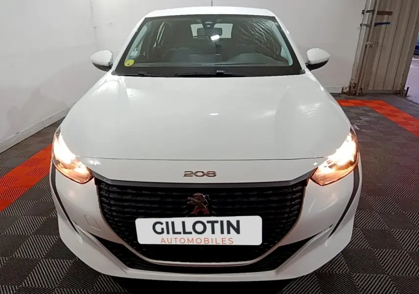 Vue frontale d'une Peugeot 208 blanche avec phares allumés et calandre noire distinctive dans un garage.