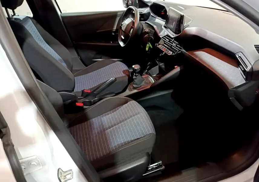Vue intérieure côté conducteur de la Peugeot 208 blanche, montrant les sièges tissu à motifs et la console avec écran tactile.