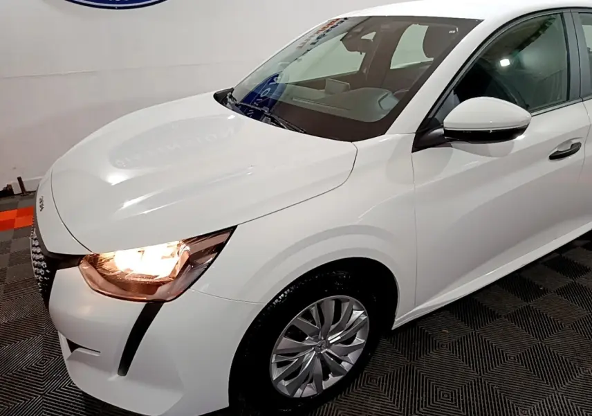 Vue 3/4 avant droit d'une Peugeot 208 blanche avec phares allumés et jantes alliage visibles.