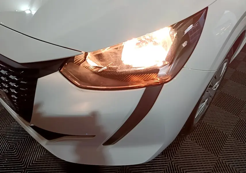 Gros plan sur le phare avant droit allumé d'une Peugeot 208 blanche, mettant en valeur la calandre et la jante.