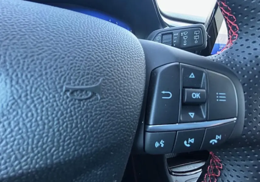 Gros plan sur le volant perforé avec surpiqûres rouges du Ford Puma gris Magnetic, montrant les commandes multifonctions.