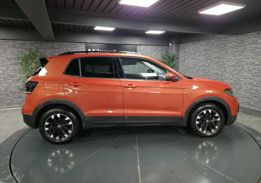 Profil droit d'un Volkswagen T-Cross orange métallisé 2021 avec jantes alliage noires et toit noir en intérieur showroom.