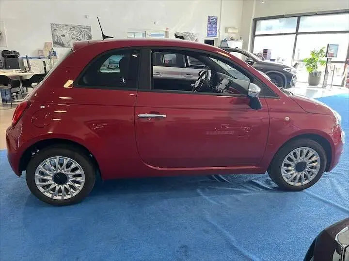 Fiat 500 rouge vue de profil côté gauche dans un showroom, avec jantes alliage et intérieur noir visible.