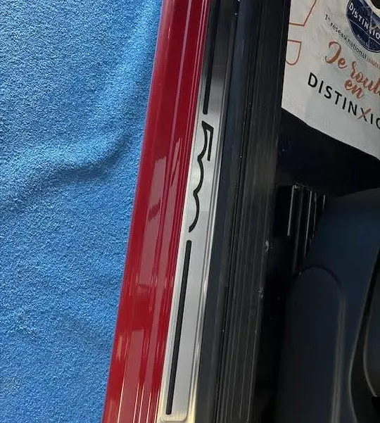 Gros plan sur le seuil de porte côté gauche d'une Fiat 500 rouge 2023 avec logo 500 visible sur fond bleu.