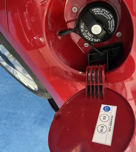 Gros plan sur la trappe à carburant rouge ouverte d'une Fiat 500 2023, avec bouchon essence et étiquette E5 E10 visible.