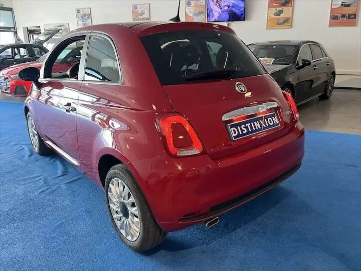 Vue 3/4 arrière droite d'une FIAT 500 rouge brillante exposée en showroom sur sol bleu, avec plaque Distinxion visible.