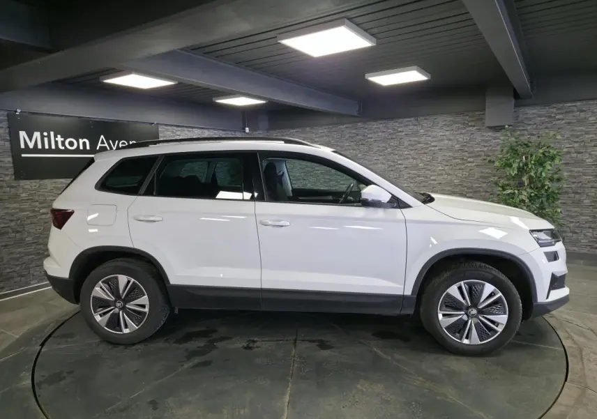 Vue de profil côté gauche d'un Skoda Karoq blanc 2023 avec jantes alliage et barres de toit noires en intérieur showroom.