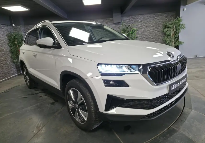 Vue 3/4 avant droit d'un Skoda Karoq blanc avec calandre noire et phares allumés en intérieur showroom.