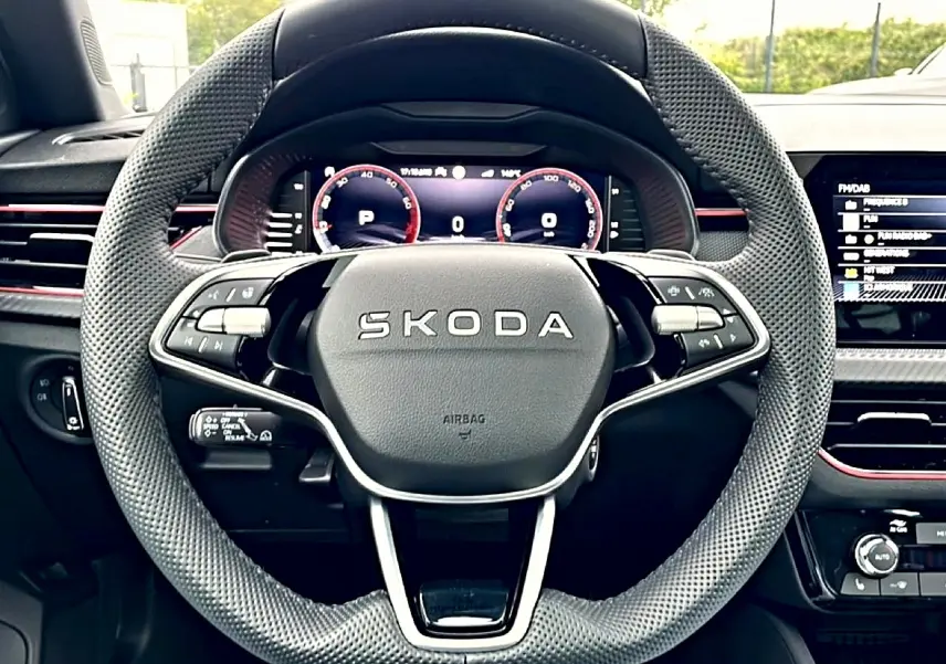 Vue rapprochée du volant cuir perforé SKODA avec tableau de bord numérique et inserts rouges sur fond noir.