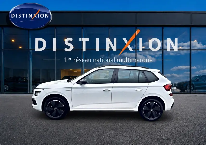 Profil droit d'un Skoda Kamiq blanc nacré 2026 avec jantes noires et toit panoramique en verre, devant une vitrine Distinxion.