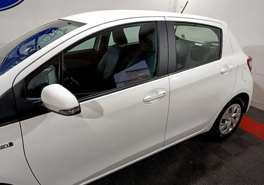 Profil côté gauche d'une Toyota Yaris blanche hybride 2019 en intérieur, mettant en valeur ses lignes compactes et ses rétroviseurs intégrés.