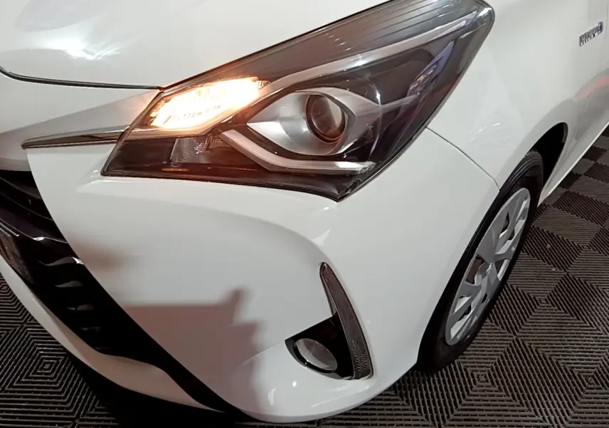 Gros plan sur l'avant droit blanc d'une Toyota Yaris hybride 2019, mettant en valeur le phare allumé et le feu antibrouillard.
