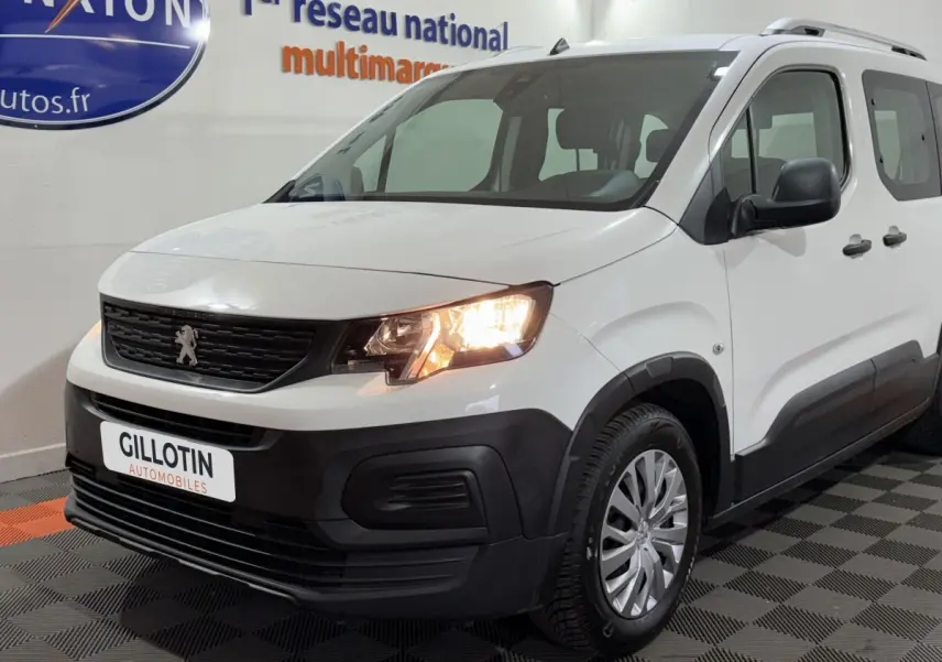 Vue 3/4 avant droit d’un Peugeot Rifter blanc de 2019 avec phares allumés et protections noires sur les bas de caisse.