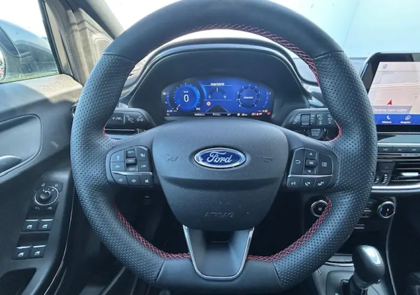 Vue intérieure centrée sur le volant noir à surpiqûres rouges du Ford Puma gris Solar avec tableau de bord numérique bleu.
