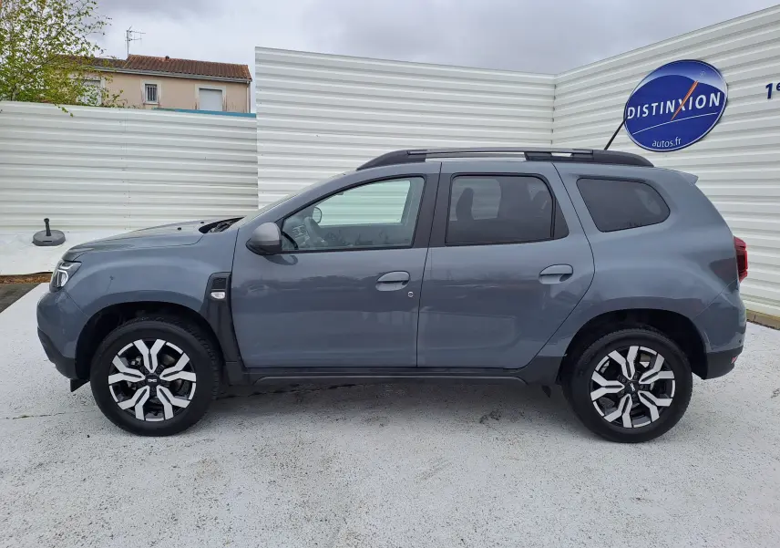 Vue de profil côté gauche d'un Dacia Duster gris urbain 2024 avec jantes bi-ton et barres de toit noires.
