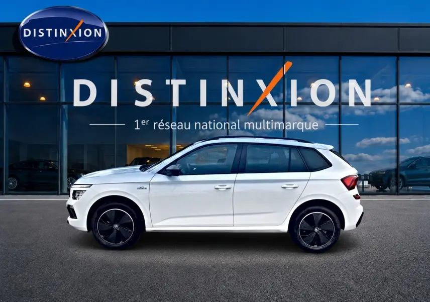 Profil côté gauche d'un Skoda Kamiq blanc nacré Monte Carlo avec jantes noires devant une vitrine Distinxion.