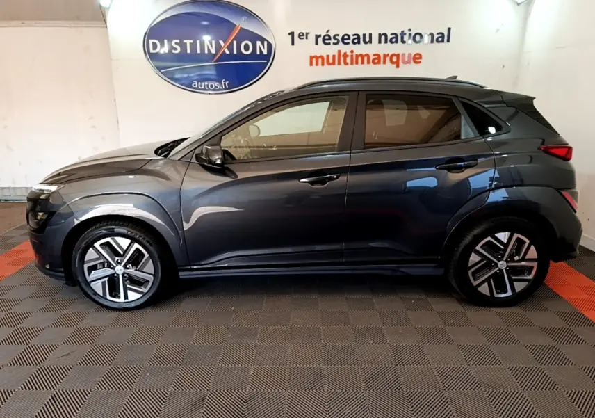 Vue de profil côté gauche d'un Hyundai Kona électrique gris foncé avec jantes distinctives en intérieur showroom.