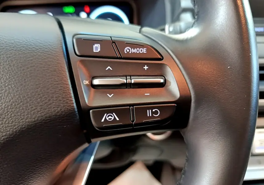 Gros plan sur les commandes du volant cuir noir du Hyundai Kona électrique, avec boutons multifonctions et régulateur de vitesse.