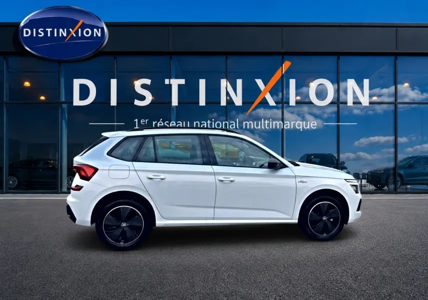 Profil droit d’un Skoda Kamiq blanc nacré 2026 avec jantes noires et toit panoramique, devant une vitrine Distinxion.
