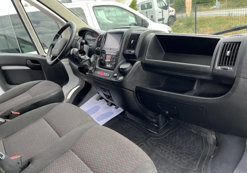 Vue intérieure côté conducteur du Peugeot Boxer Fourgon 2023, montrant tableau de bord noir et sièges tissu gris à motifs rouges.