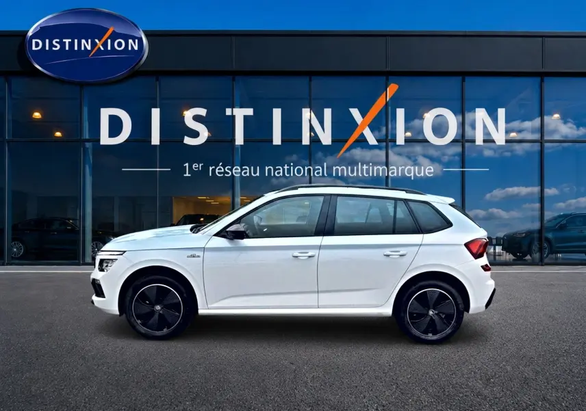 Vue latérale droite d'un Skoda Kamiq blanc nacré avec jantes noires devant un bâtiment vitré Distinxion.