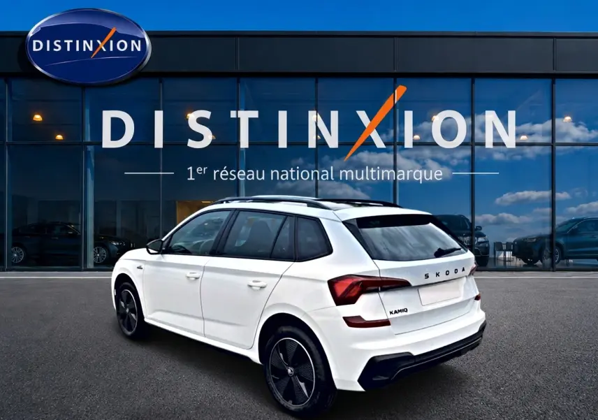 Vue 3/4 arrière droite d’un Skoda Kamiq blanc nacré avec jantes noires devant un bâtiment vitré Distinxion.