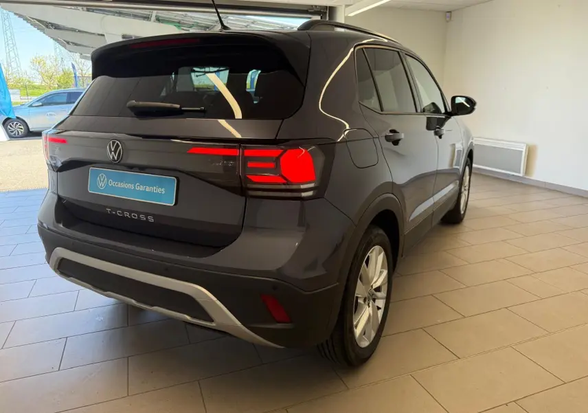 Volkswagen T-Cross gris cendre vue 3/4 arrière droit en intérieur, feux arrière allumés et logo VW visible.