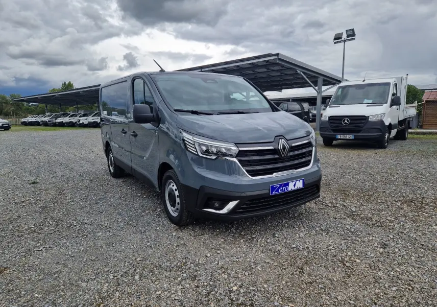 Renault Trafic Fourgon gris urbain vu en 3/4 avant droit sur un parking gravillonné avec plusieurs véhicules en arrière-plan.