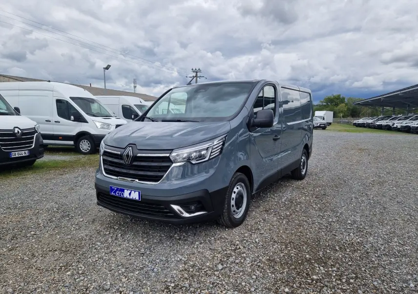 Renault Trafic Fourgon gris urbain vu en 3/4 avant droit sur un parking gravillonné, avec calandre noire et phares LED.