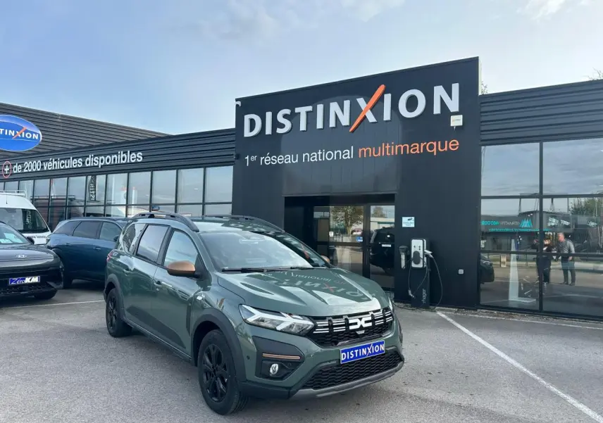 Vue 3/4 avant d'un Dacia Jogger kaki lichen métal avec calandre noire et jantes noires devant un concessionnaire Distinxion.