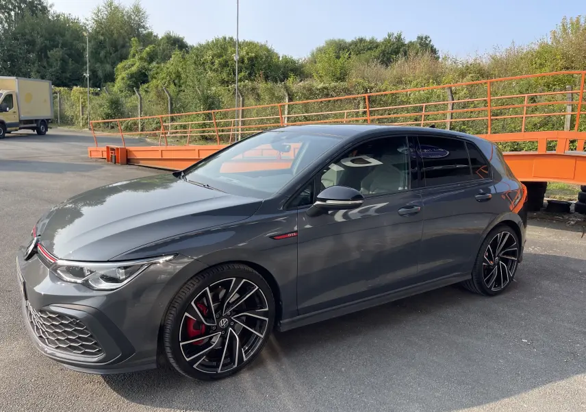 Volkswagen Golf VIII GTI gris vue 3/4 avant droit, jantes noires à détails rouges et calandre sport avec liseré rouge.