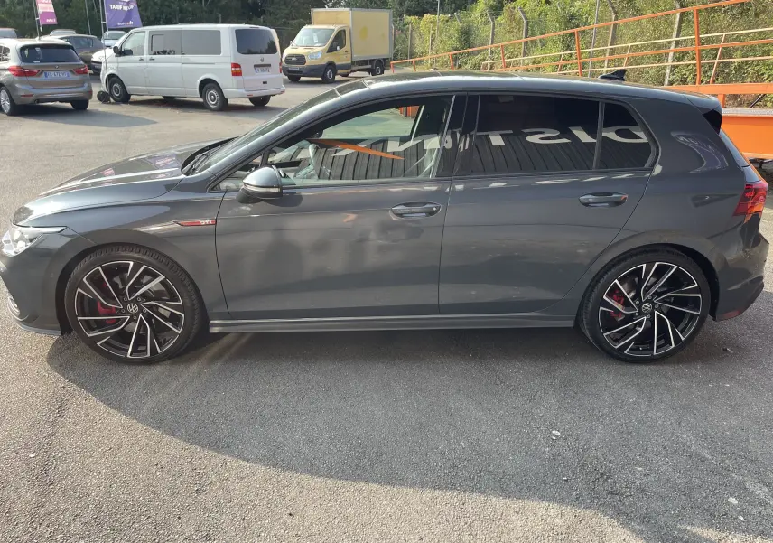 Profil côté gauche d'une Volkswagen Golf VIII GTI gris métallisé avec jantes noires et rouges sportives, en extérieur.