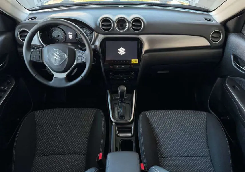 Intérieur du Suzuki Vitara 2025 vu de face, tableau de bord noir avec écran central tactile et sièges tissu foncé.