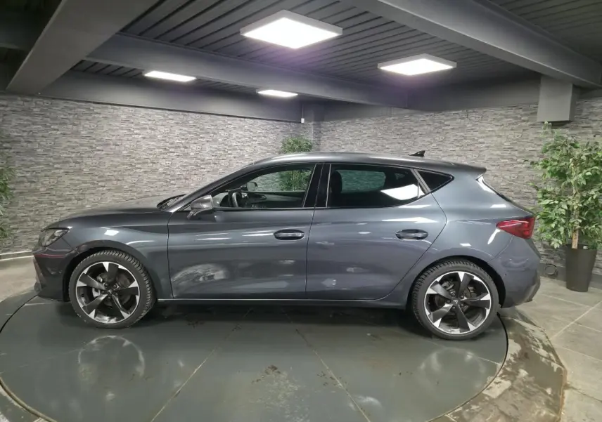 Vue de profil côté gauche d'une CUPRA Leon gris magnétique avec jantes alliage 18'' dans un showroom intérieur.