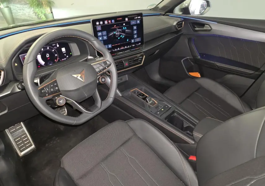 Intérieur avant de la CUPRA Leon 2025 gris, vue côté conducteur avec volant cuir, écran tactile et éclairage d’ambiance bleu.