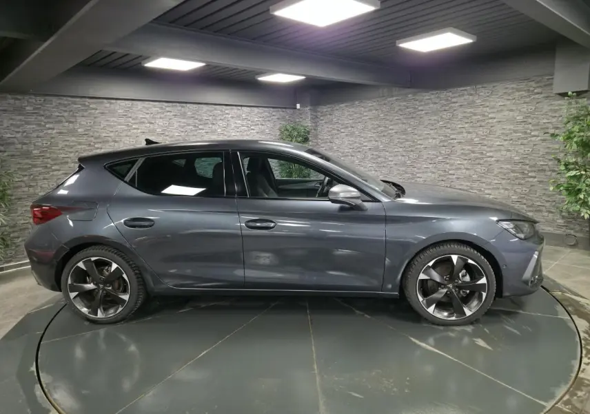 Vue de profil côté gauche d'une CUPRA Leon gris magnétique avec jantes alliage 18 pouces dans un showroom intérieur.