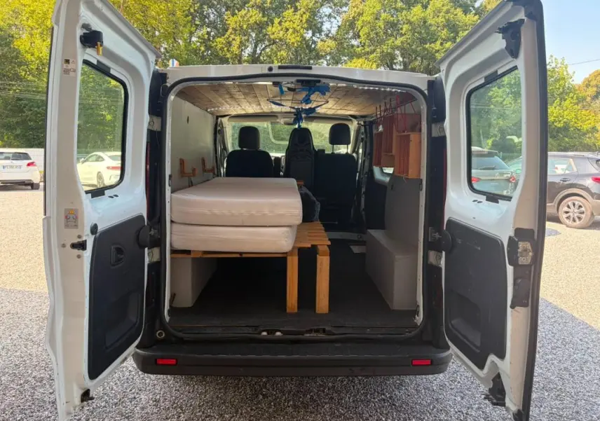Vue arrière du Renault Trafic Fourgon blanc 2018, portes ouvertes montrant l’aménagement intérieur avec lit et rangements en bois.