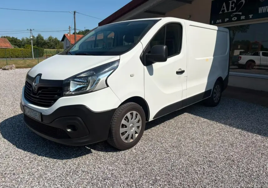 Renault Trafic Fourgon blanc en 3/4 avant droit, avec pare-chocs noir et jantes acier, stationné sur gravier.