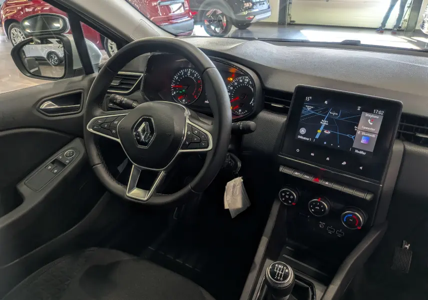 Vue intérieure côté conducteur de la Renault Clio V gris platine, avec tableau de bord digital et levier de vitesse manuel.