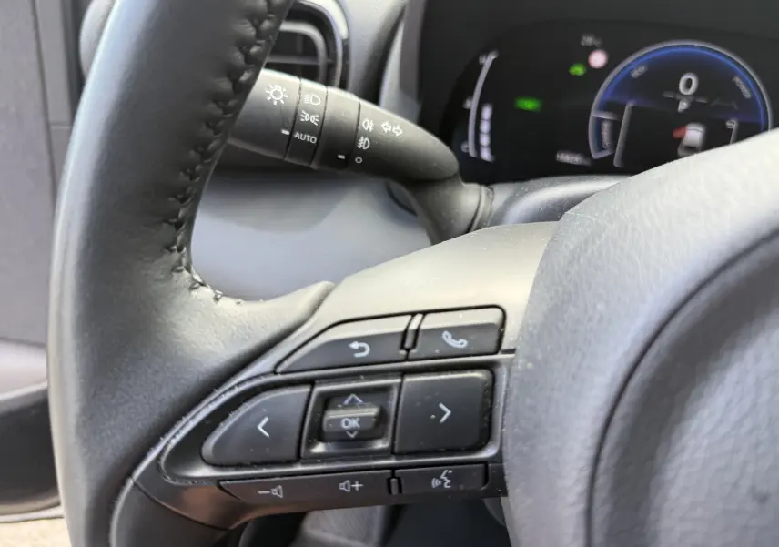 Gros plan sur le volant noir de la Toyota Yaris Cross 130 CH Design 2024, avec commandes intégrées et tableau de bord numérique en arrière-plan.