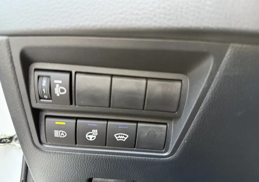 Gros plan sur les boutons de commande intérieurs noirs du Toyota Yaris Cross blanc, avec réglage des phares et chauffage du volant.