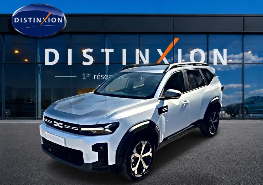 Dacia Bigster blanc en 3/4 avant droit, avec jantes alu et barres de toit visibles devant un showroom Distinxion.