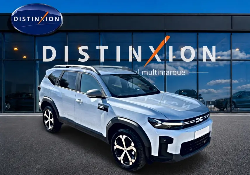 SUV Dacia Bigster blanc en 3/4 avant droit, avec barres de toit et jantes alu distinctives, devant une vitrine multimarque.